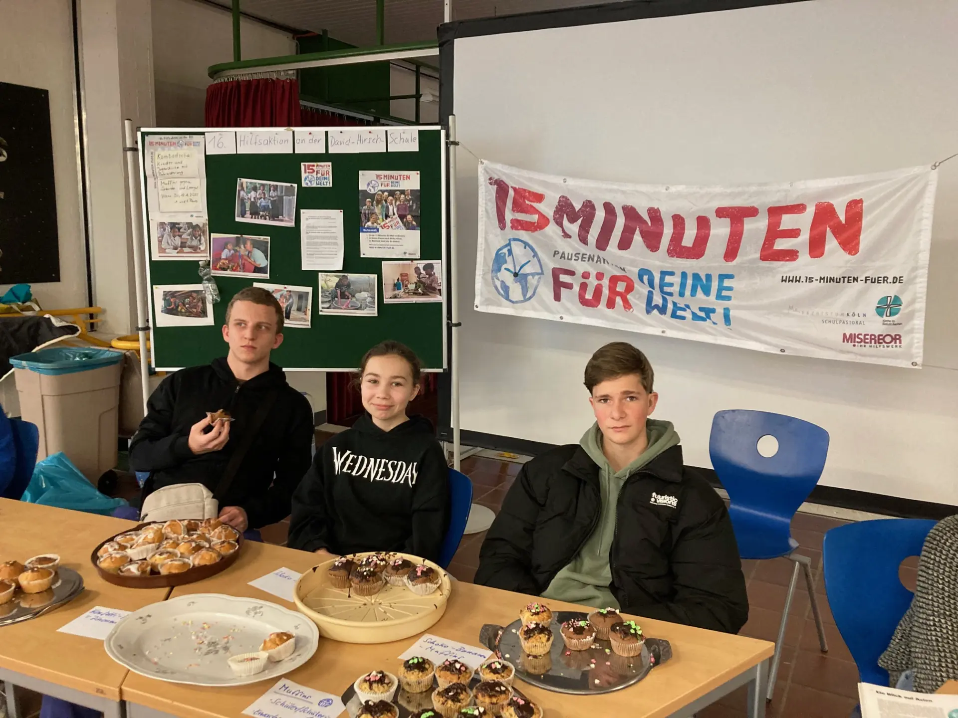 drei schüler sitzen bei einem kuchenverkauf hinter einem tisch mit verschiedenen kuchen