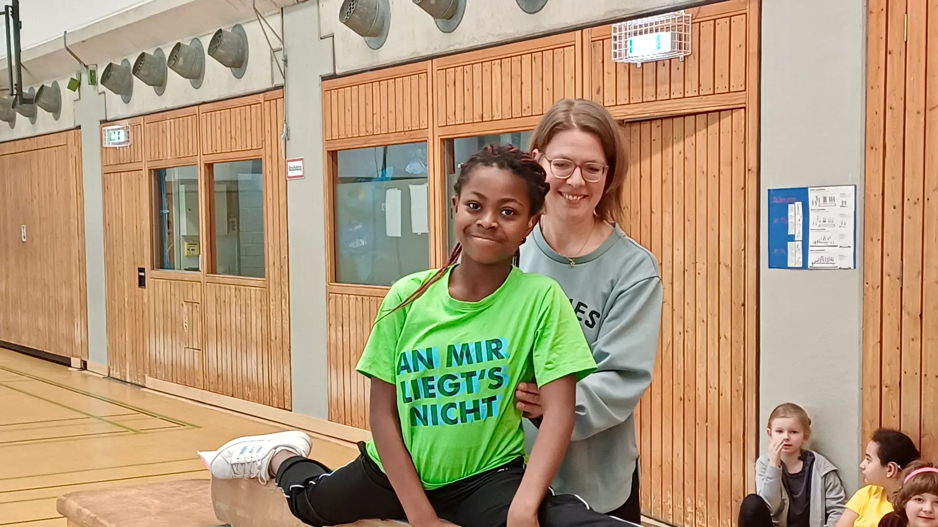 ein mädchen zeigt auf dem schwebebalken mit hilfestellung durch eine lehrerin einen spagat