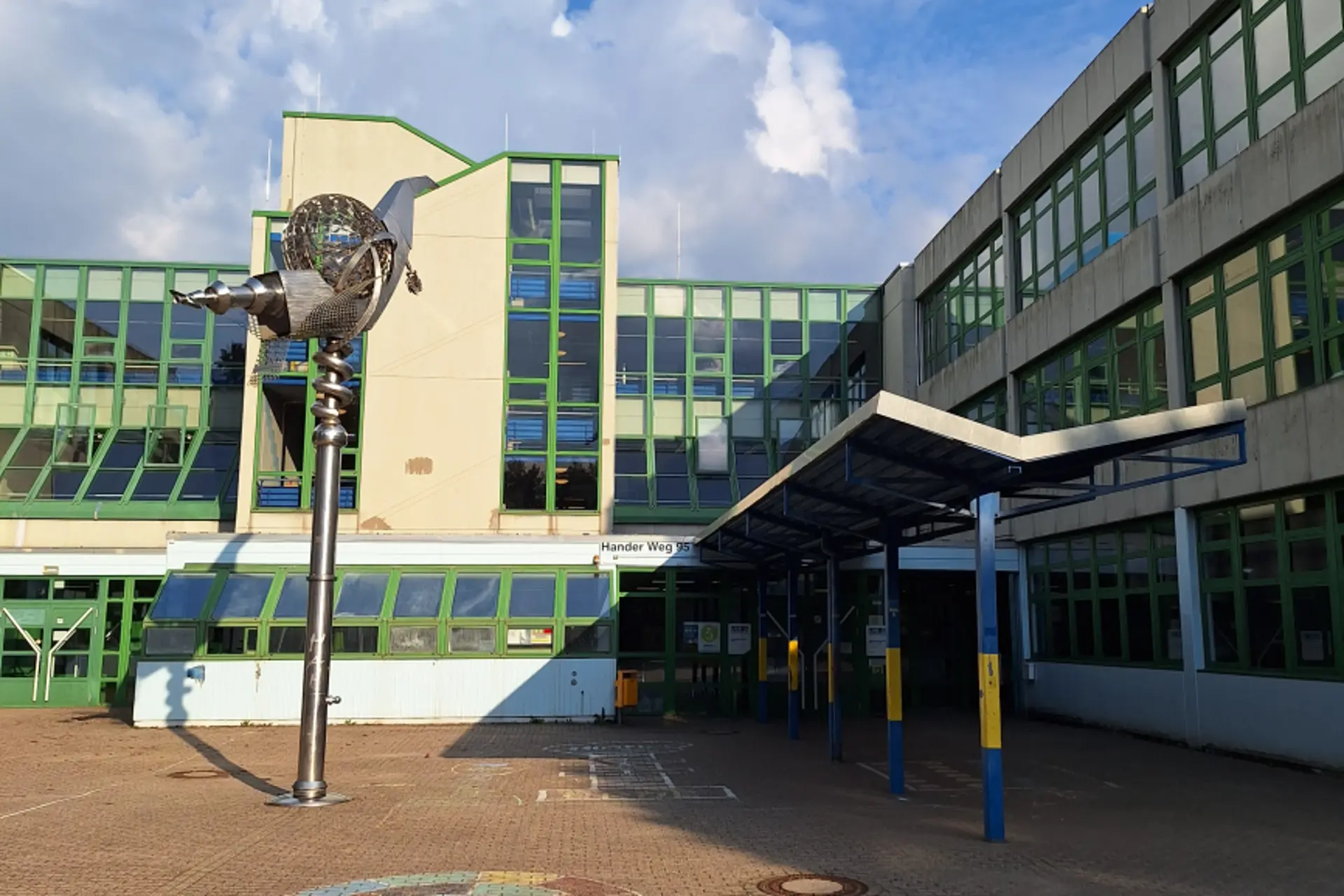 Blick auf das Schulgebäude mit einer Fensterfront mit grünen Rahmen. Im Vordergrund ist ein hohe, abstrakte Metallskulptur zu sehen.