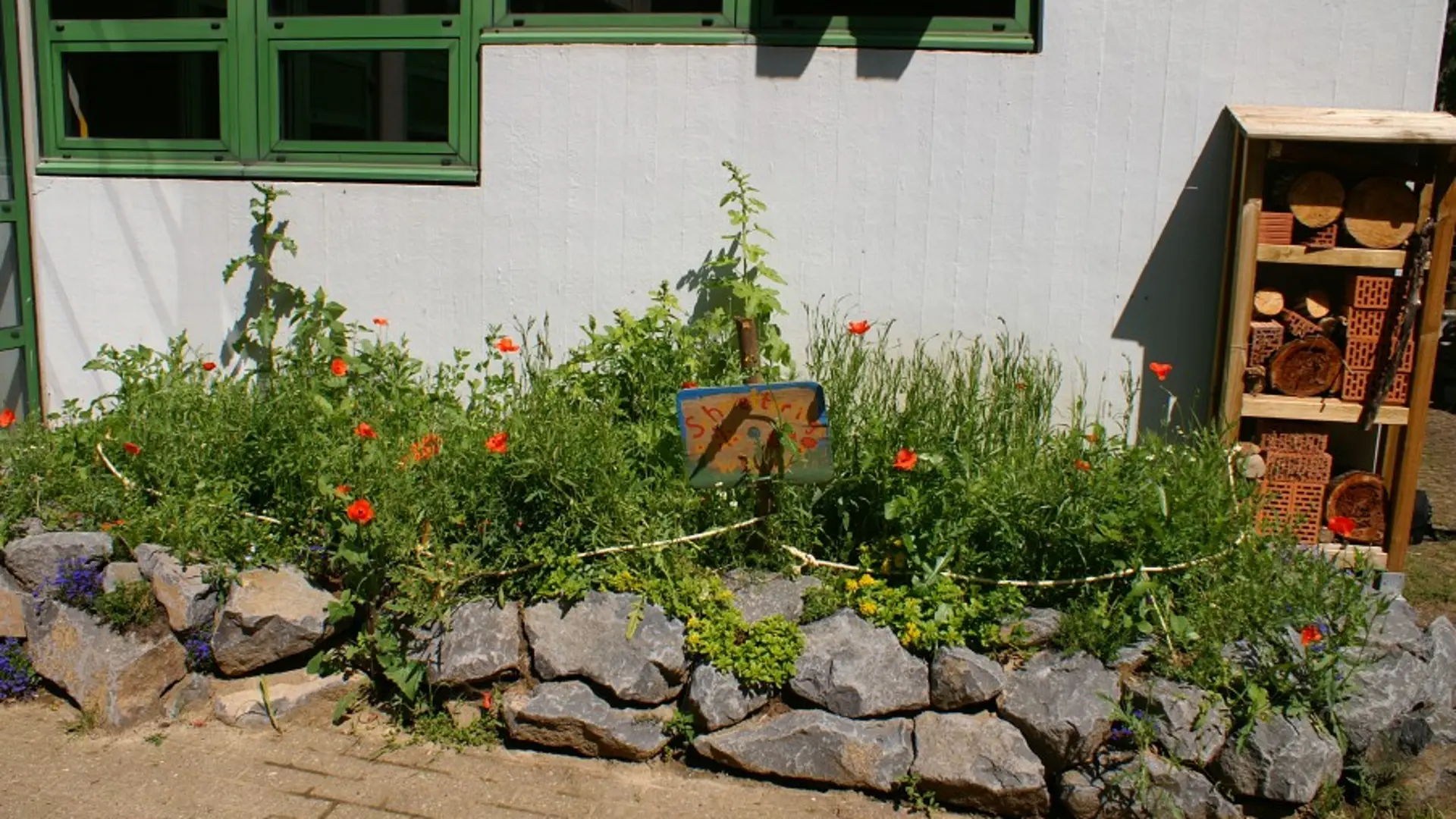 Blick auf ein Beet vor einer weißen Hauswand. Es ist mit grauen Bruchsteinen eingefasst. Im Hintergrund steht ein hohes Insektenhotel.