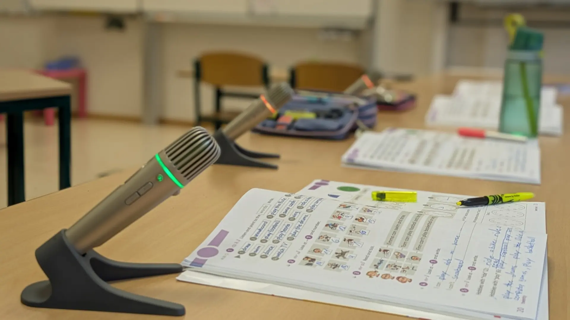 Auf Tischen im Klassenzimmer liegen Arbeitshefte und Stifte und jeweils ein Mikrofon. 