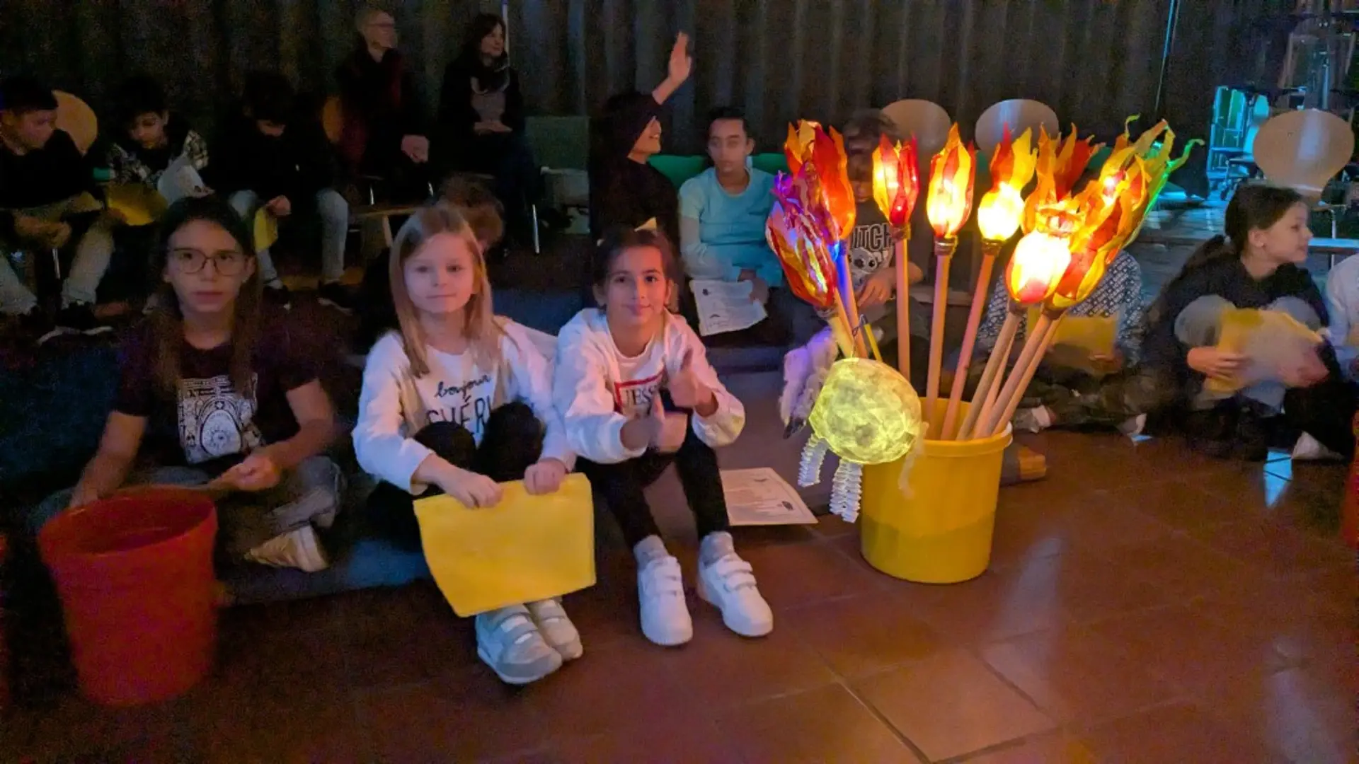 Blick in einen abgedunkelten Raum. Dort sitzen Kinder auf dem Boden. In einem Eimer stecken leuchtenden Stablaternen in Fackelform.