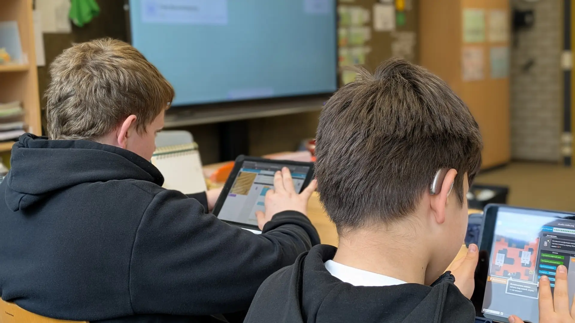 Zwei Schüler von hinten, einer trägt ein Hörgerät. Sie sitzen an Tischen im Klassenraum und arbeiten mit Tablets.