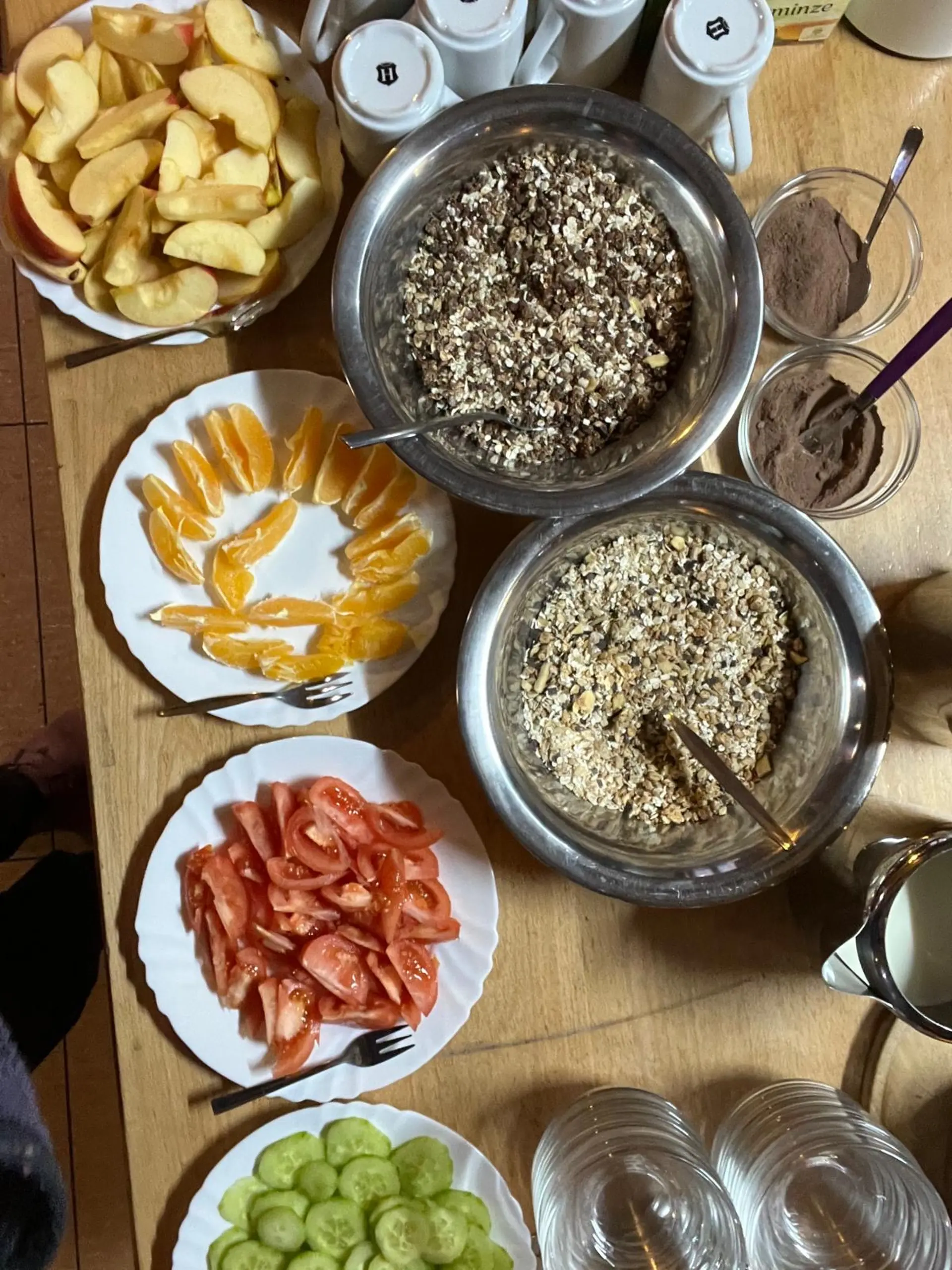 Zu sehen ist eine Frühstückstisch von oben fotografiert. Es gibt Teller mit geschnittenen Äpfeln, Orangen, Tomaten und Gurken, sowie zwei große Schüsseln mit Müsli.