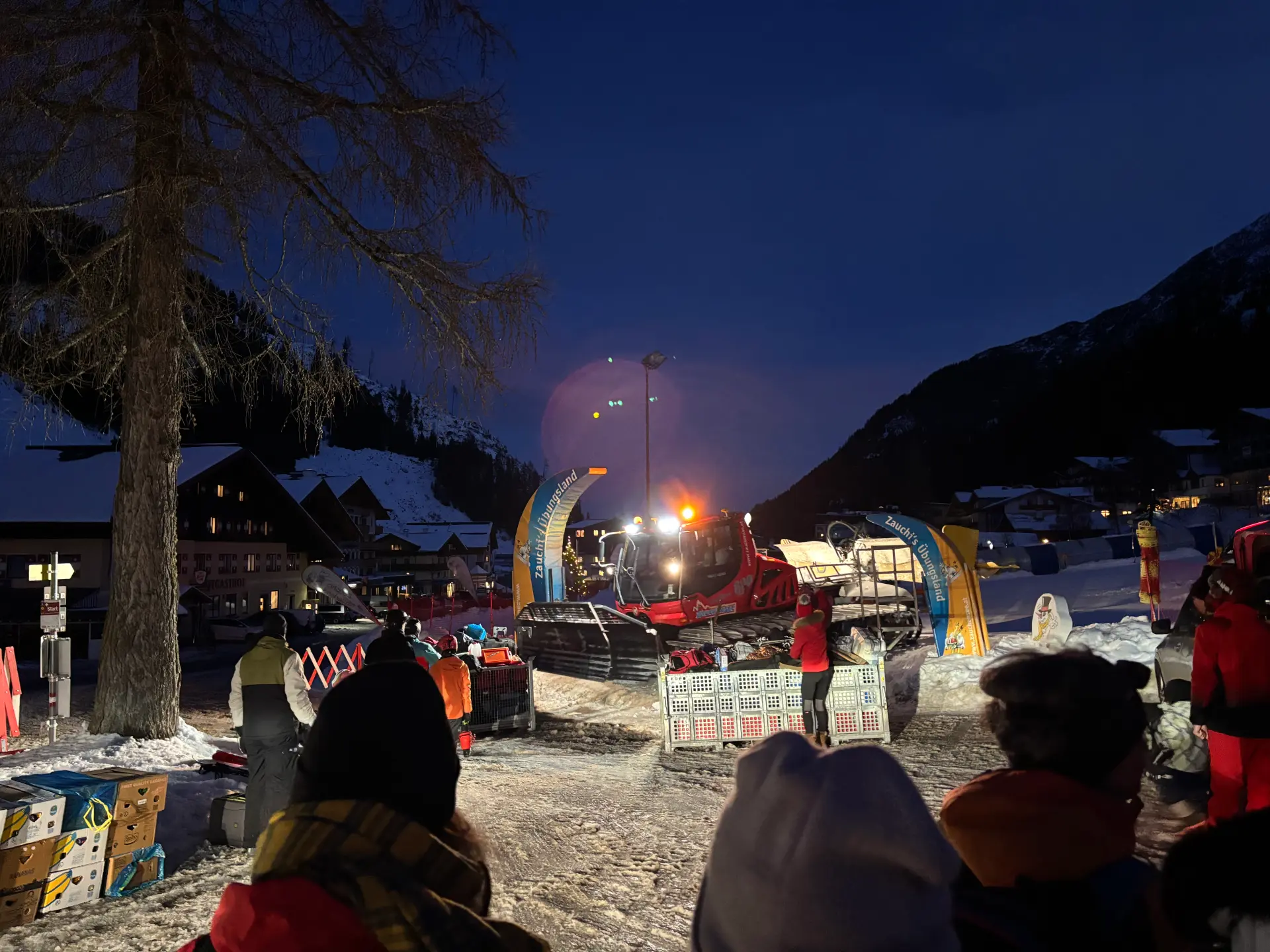 Eine Foto bei Nacht über mehrere Menschen hinweg. Zu sehen ist eine Schneeraupe.