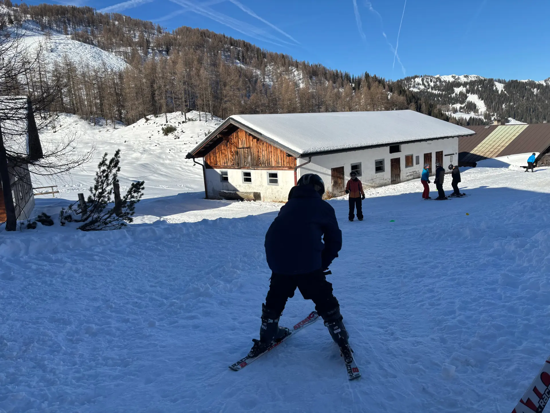 Ein Schüler macht auf Skiern einfache Bremsübungen vor einer Holzhütte in einer Winterlandschaft.