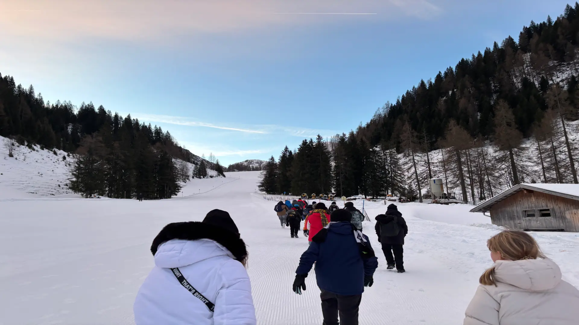 Eine Hand voll Menschen läuft eine flache Skipiste bei Tag hoch. Im Hintergrund ist eine kleine Hütte zu erkennen.