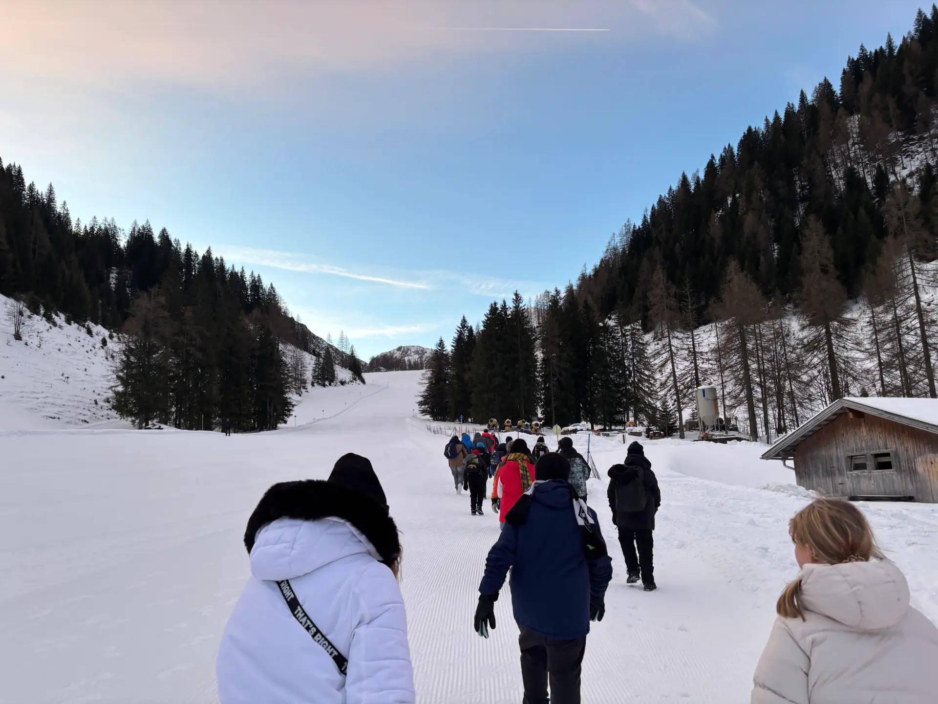 Eine Hand voll Menschen läuft eine flache Skipiste bei Tag hoch. Im Hintergrund ist eine kleine Hütte zu erkennen.