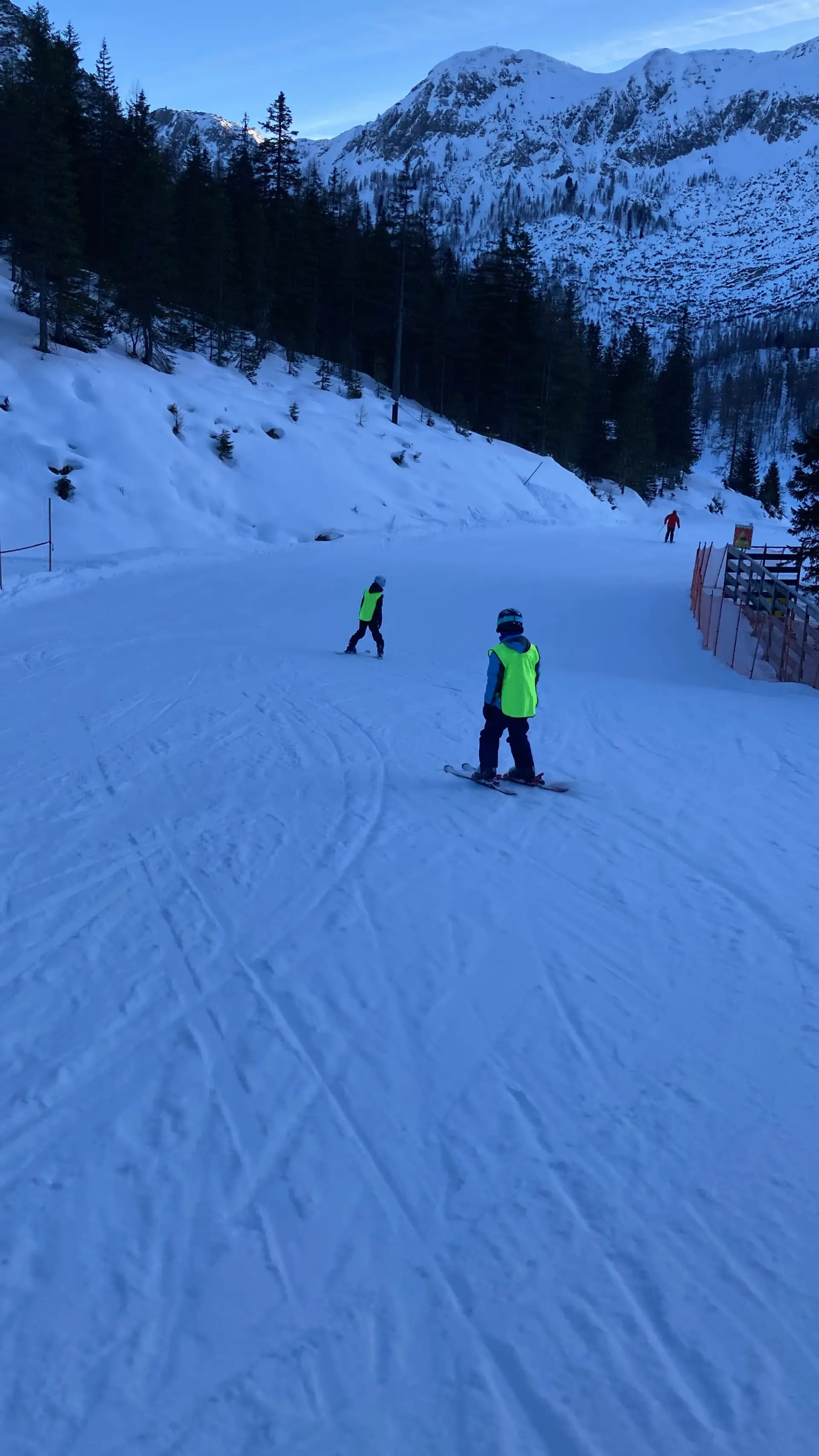 Zwei Schüler in Skikleidung und mit gelber Warnweste fahren auf Skiern eine einfache Piste hinunter. Die Schüler sind von hinten zu sehen.